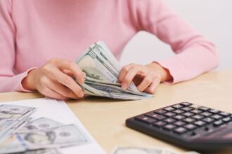 A woman sitting at a desk in her home office, counting cash for her bills and using a calculator.