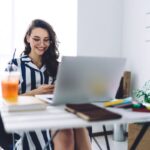 A remote freelancer is working from her home office at a desk, using a laptop and a smart phone.