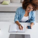 A woman writing in a notebook and working on her LinkedIn profile at a home office desk.