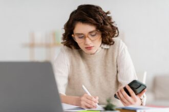 A woman at home sitting at her home office desk, using a laptop and cell phone.