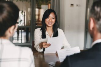 A woman sitting down in an office across from two people for an interview. She's handing them her resume.