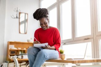 A woman working from home, sitting near a window, writing in a notebook.