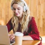 A woman working at home from her laptop, wearing headphones, and drinking coffee.