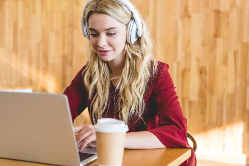 A woman working at home from her laptop, wearing headphones, and drinking coffee.