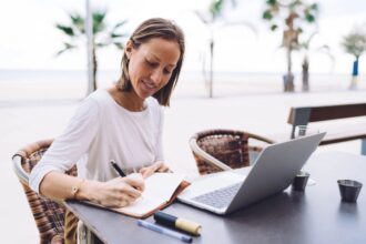 A woman working remotely on a laptop from a cafe table, with a beach and palm trees behind her.