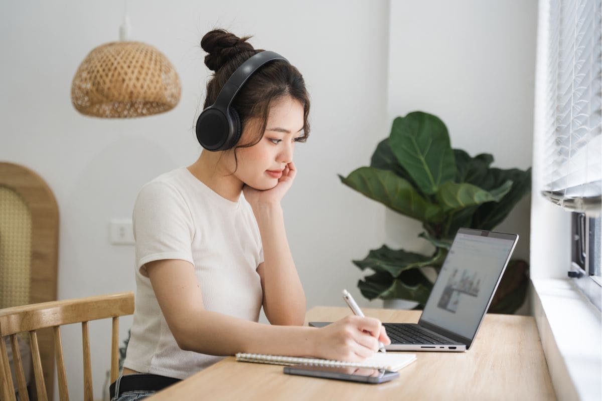 A woman sitting at her kitchen table, wearing headphones and working on a laptop.