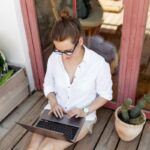 Woman sitting on a wooden patio next to potted plants, using a laptop to job hunt for remote jobs.