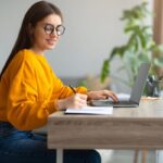 A woman with ADHD working at home using a laptop and writing in a notebook.
