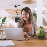 A digital nomad, working remotely on her laptop from a cafe in a tropical location, drinking from a coconut.