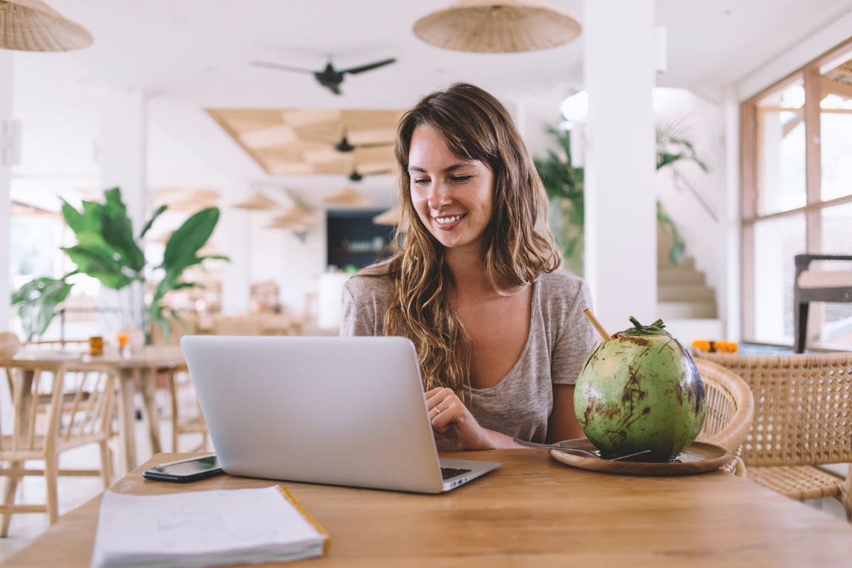 A digital nomad, working remotely on her laptop from a cafe in a tropical location, drinking from a coconut.