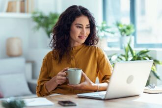 A woman working from her home office desk, holding a coffee cup, smiling, and reading on her laptop.