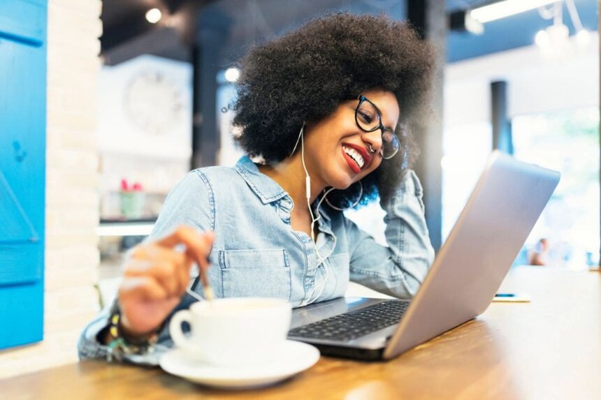 A woman working remotely from a cafe, using a laptop and drinking coffee.
