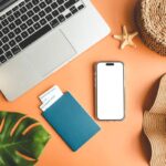 A work from home travel planner's desk, with a laptop, passport, cell phone, and sun hat.