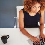Overhead view of a woman working at her home office desk, drinking coffee and typing on a keyboard.