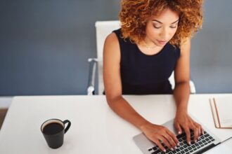 Overhead view of a woman working at her home office desk, drinking coffee and typing on a keyboard.