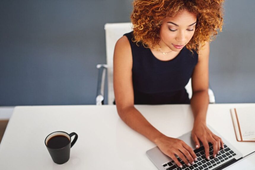 Overhead view of a woman working at her home office desk, drinking coffee and typing on a keyboard.