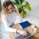 A young woman is working remotely in a cafe on a laptop and talking on the phone.